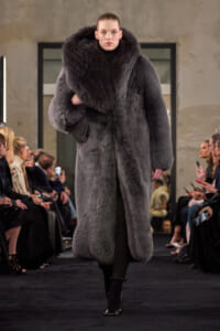 Model on a fashion runway wearing a voluminous grey fur coat with a high collar, black leggings, and black heeled boots.
