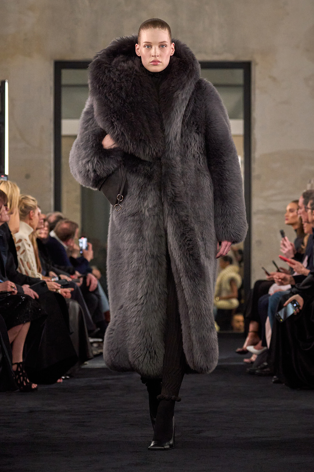 Model on a fashion runway wearing a voluminous grey fur coat with a high collar, black leggings, and black heeled boots.