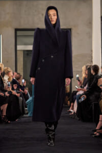 Model walking a fashion show in a navy oversized hooded coat with a wrap collar and black boots, carrying a turquoise bag.