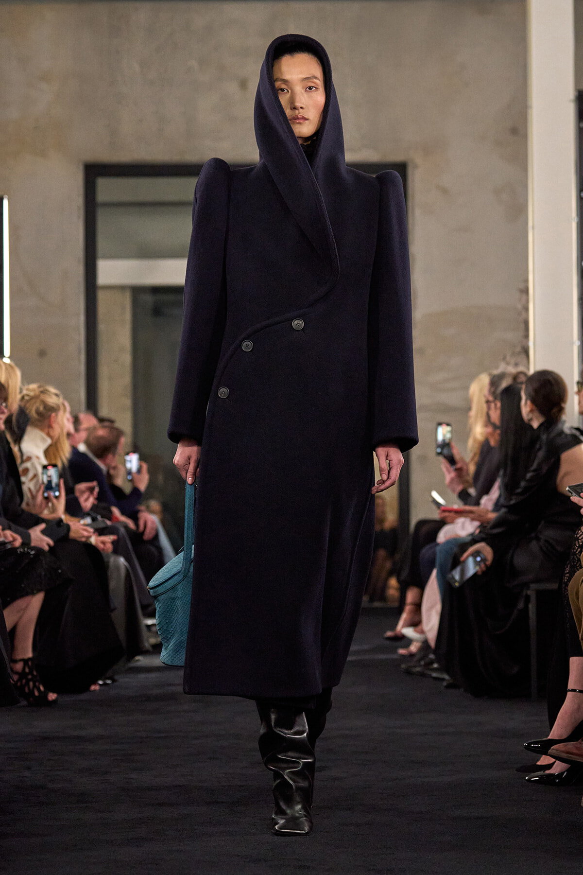 Model walking a fashion show in a navy oversized hooded coat with a wrap collar and black boots, carrying a turquoise bag.
