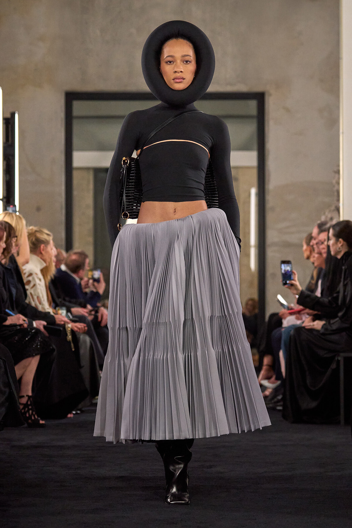 Model on a runway wearing a black hooded crop top with a circular neckline and a voluminous grey pleated skirt, with black ankle boots and a black shoulder bag.