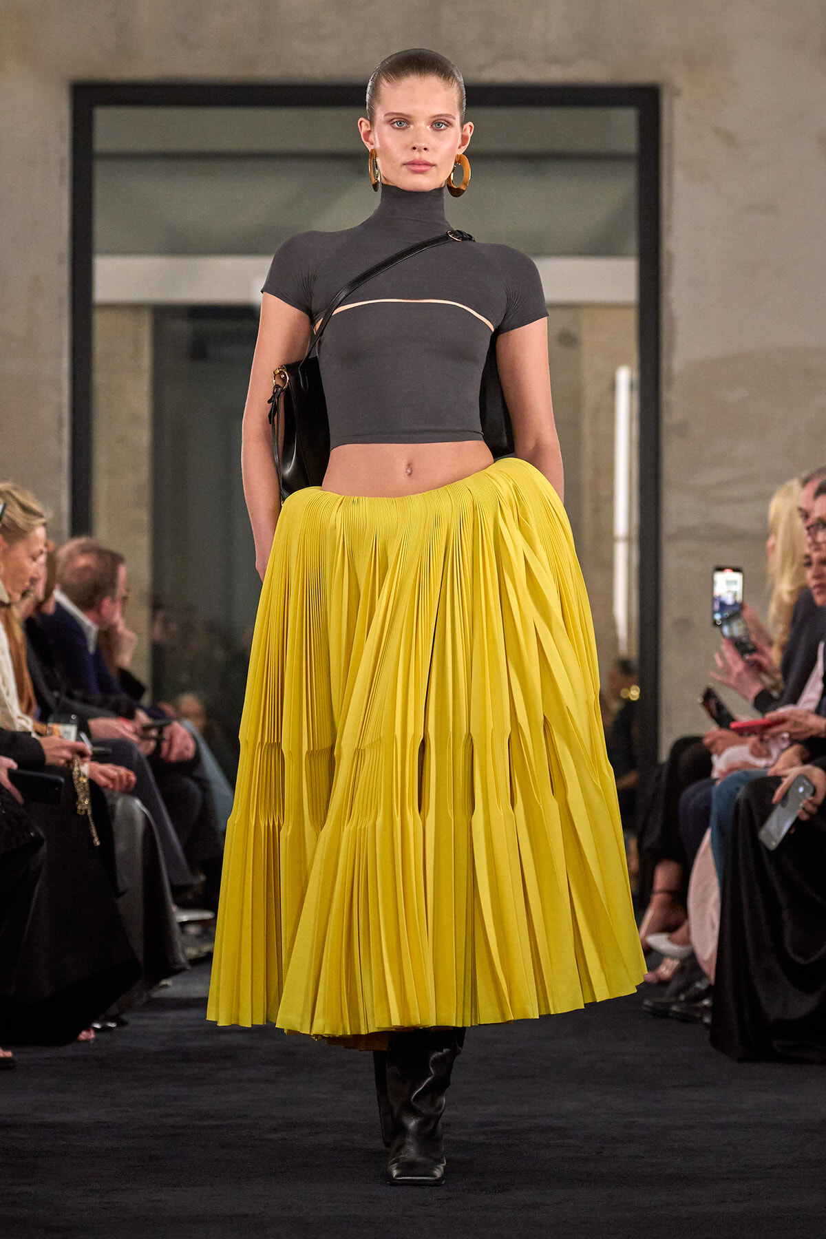 Model walks the runway in a charcoal cropped top and bright yellow pleated midi skirt, with a black bag and gold hoop earrings.—fashion show scene