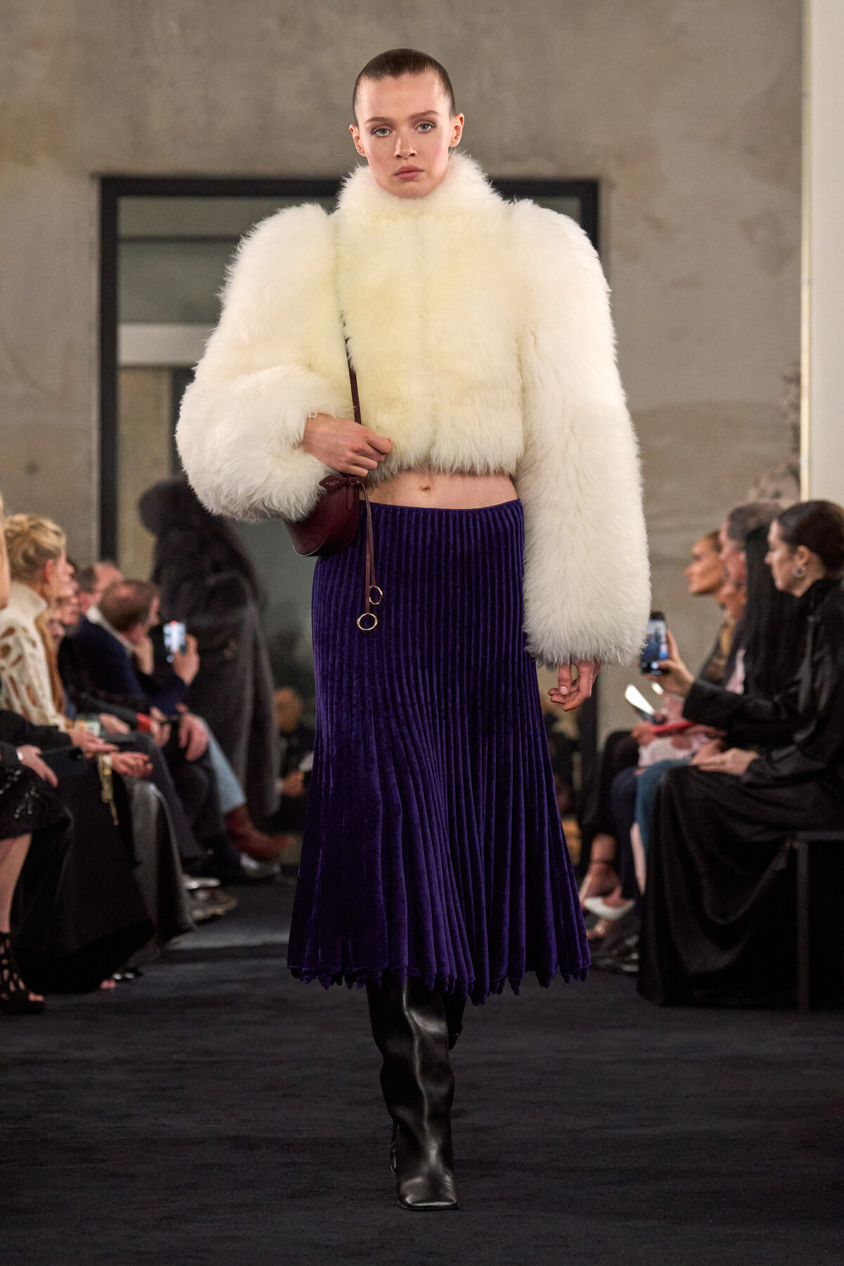 Model walks a runway in a white fur cropped jacket, a purple velvet pleated midi skirt, and black knee boots, carrying a small brown bag.