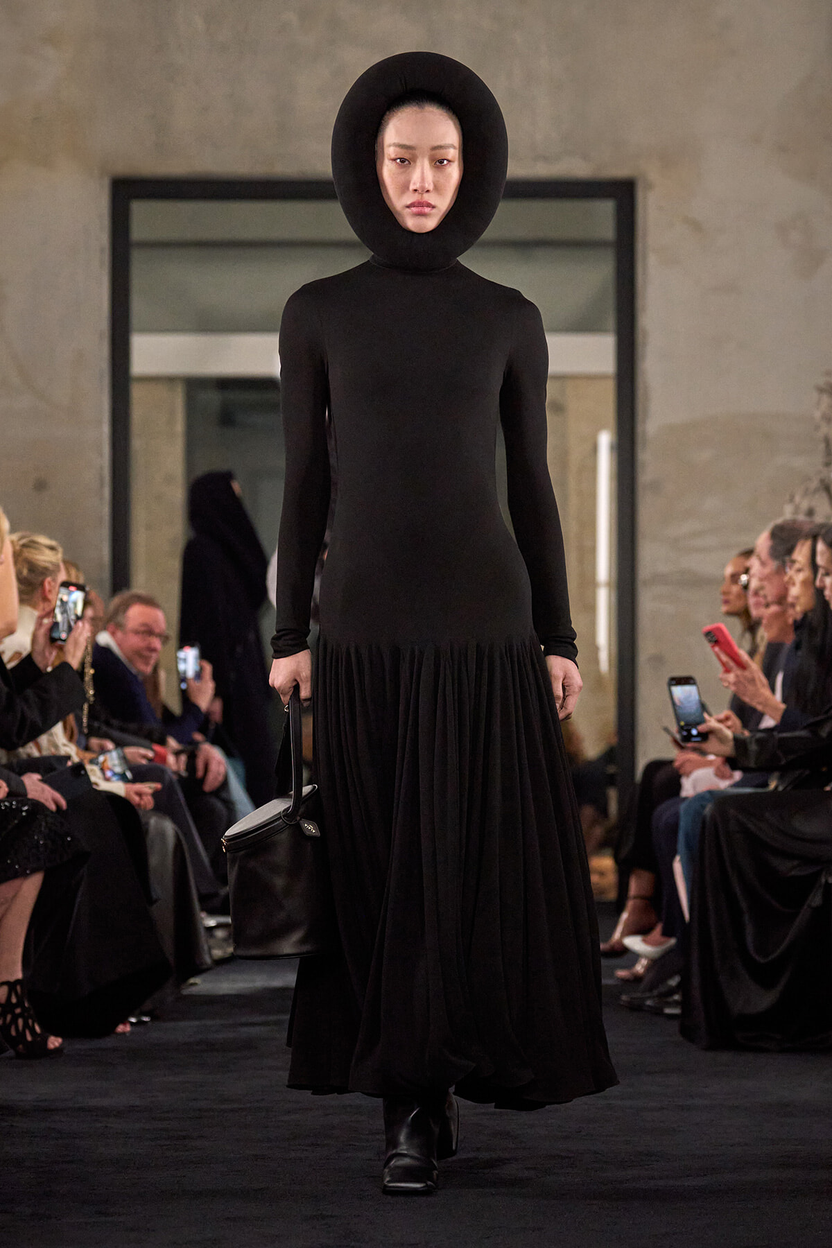 Model walks the runway in an all-black ensemble with a sculptural circular hood, fitted top and voluminous pleated skirt, carrying a black handbag.