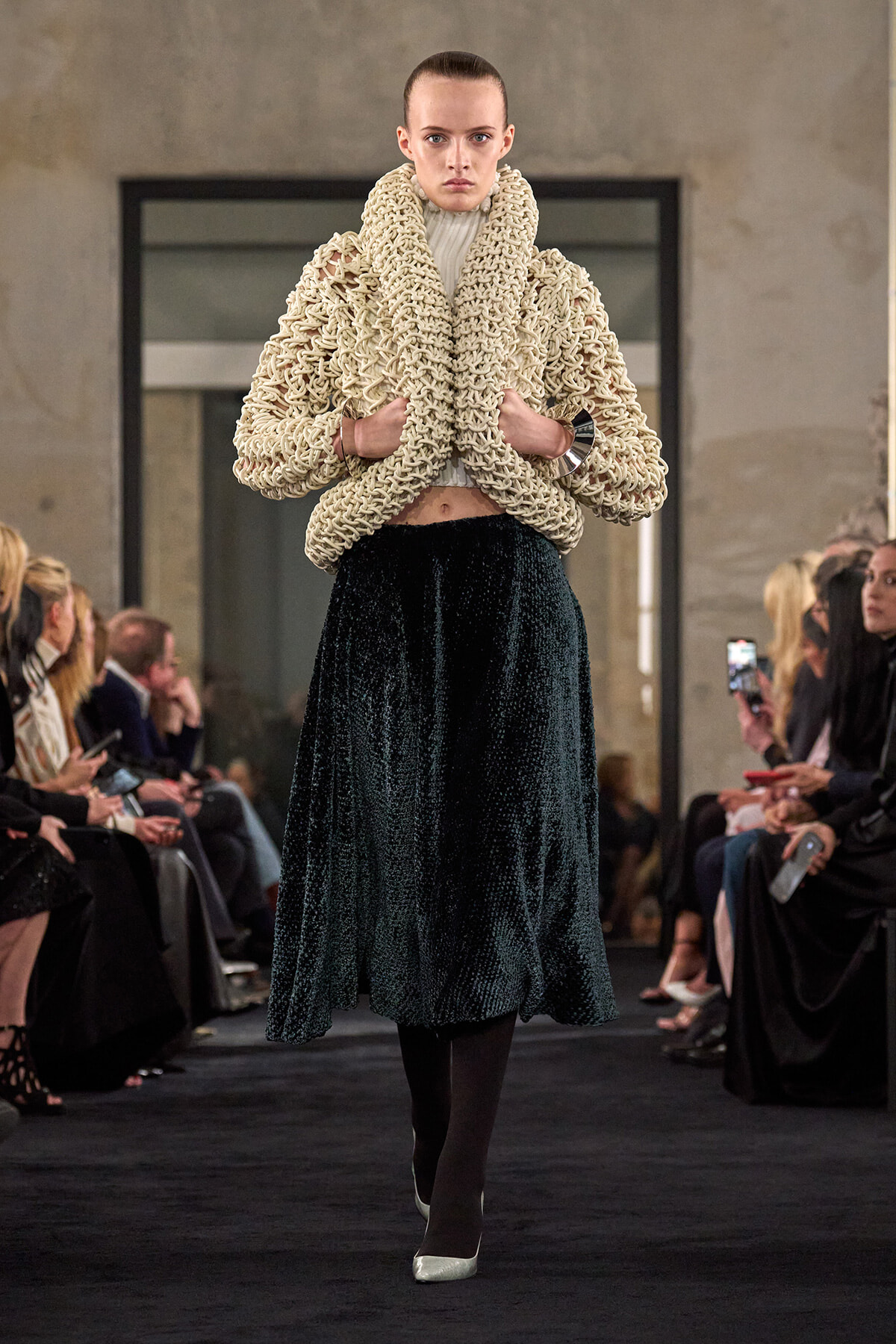 Model walks the runway in an oversized cream chunky knit coat paired with a dark velvet midi skirt.