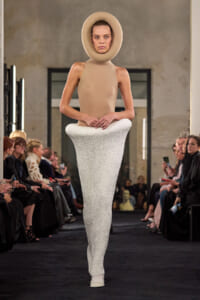 Model walks runway in an avant-garde ensemble: a circular beige headpiece, sleeveless beige top, and a long white textured skirt-enhanced silhouette; audience on both sides.