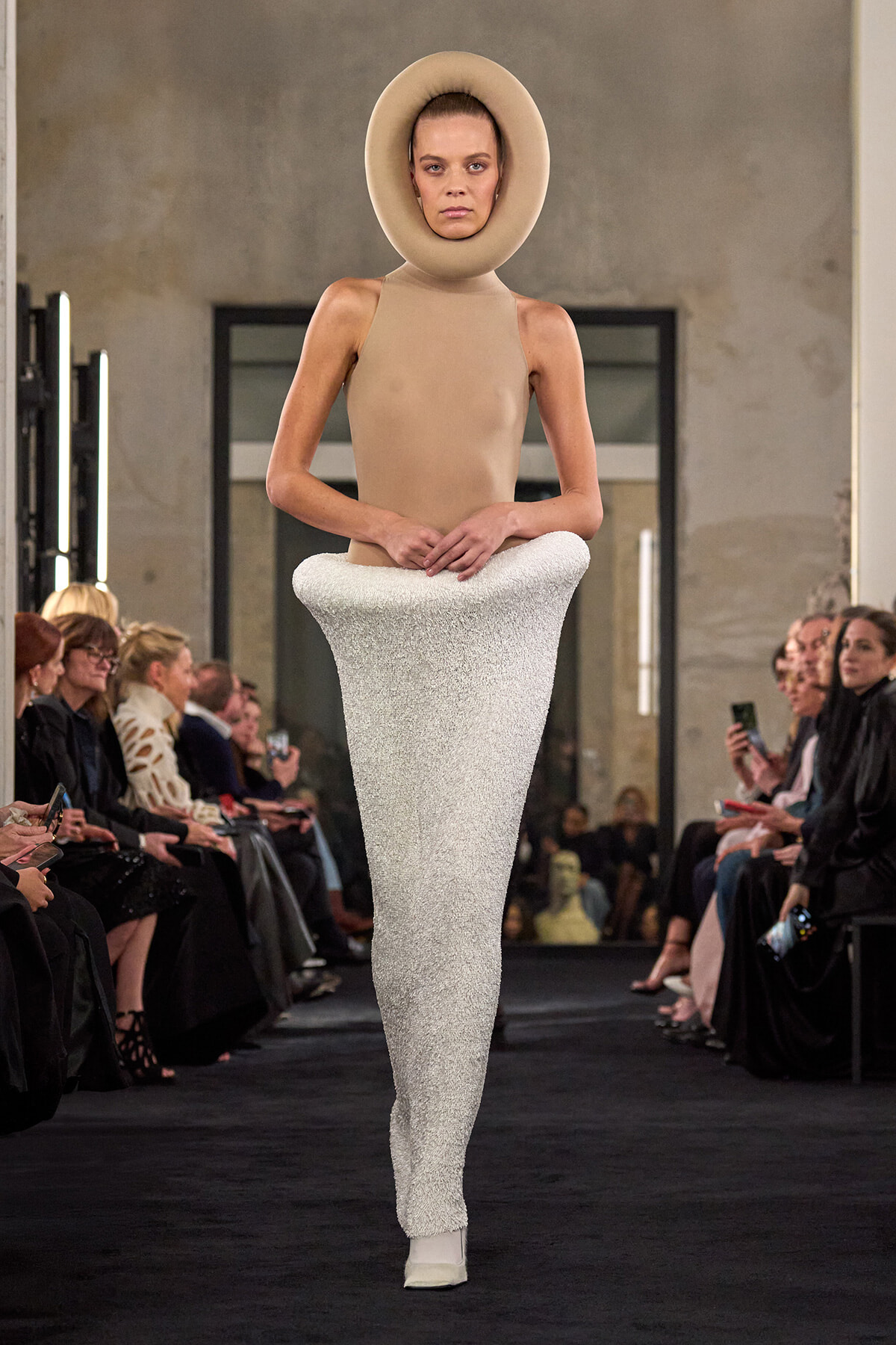 Model walks runway in an avant-garde ensemble: a circular beige headpiece, sleeveless beige top, and a long white textured skirt-enhanced silhouette; audience on both sides.