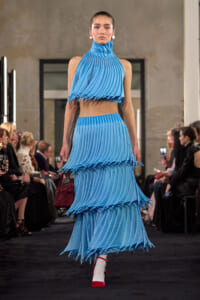 Model in cobalt blue pleated two-piece dress with a high ruffled neck on a runway, carrying a burgundy handbag.
