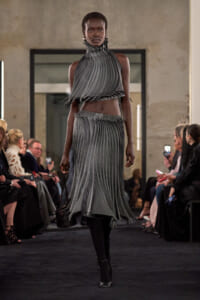 Runway model wearing a grey pleated two-piece dress with a high, ruffled collar walking the catwalk as spectators watch.