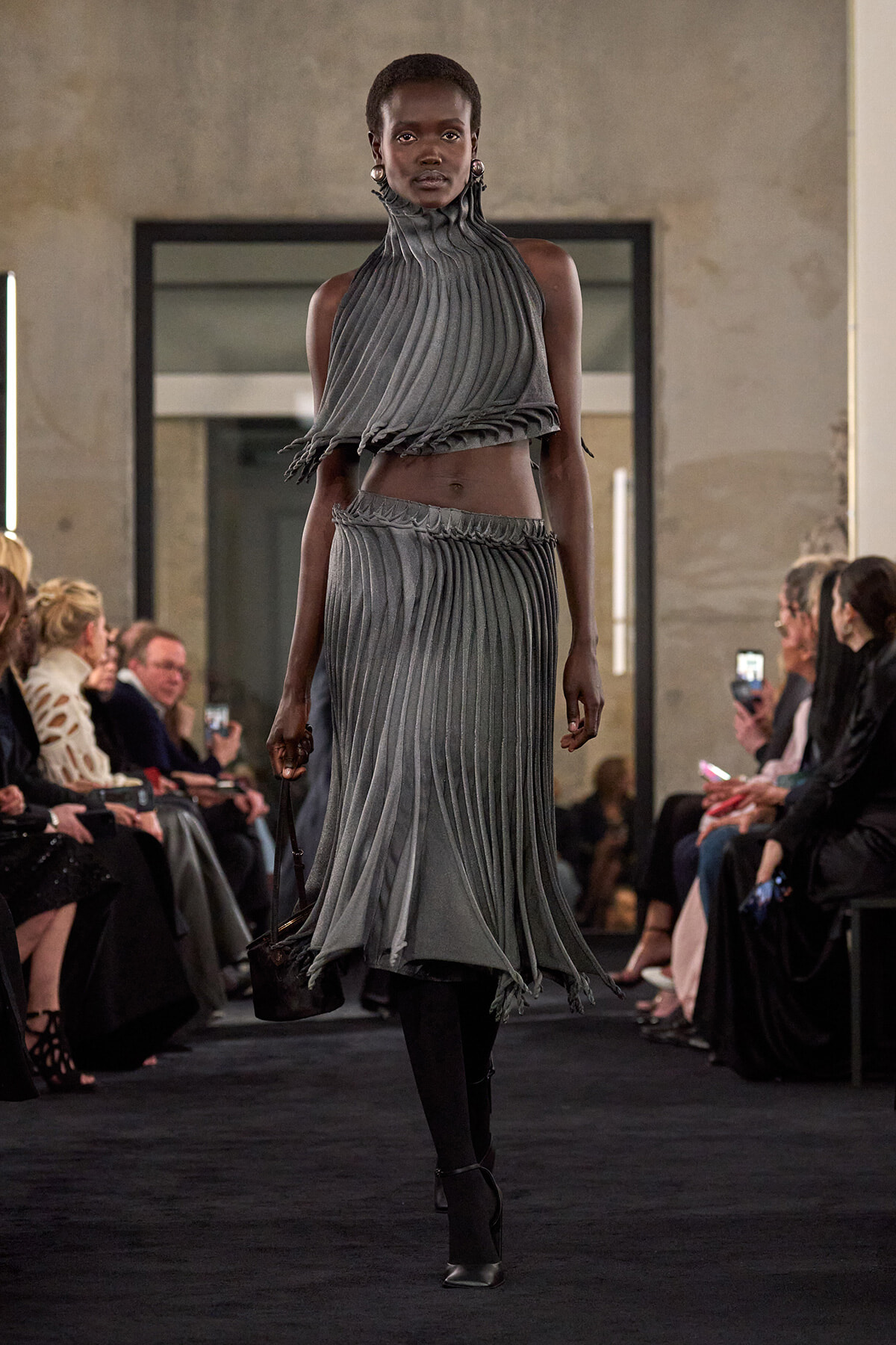 Runway model wearing a grey pleated two-piece dress with a high, ruffled collar walking the catwalk as spectators watch.