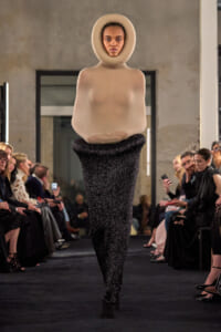 Runway model wearing a nude, sculptural bodysuit with a large circular collar around the head, paired with a glittery black bottom piece, walking the catwalk as an audience watches.