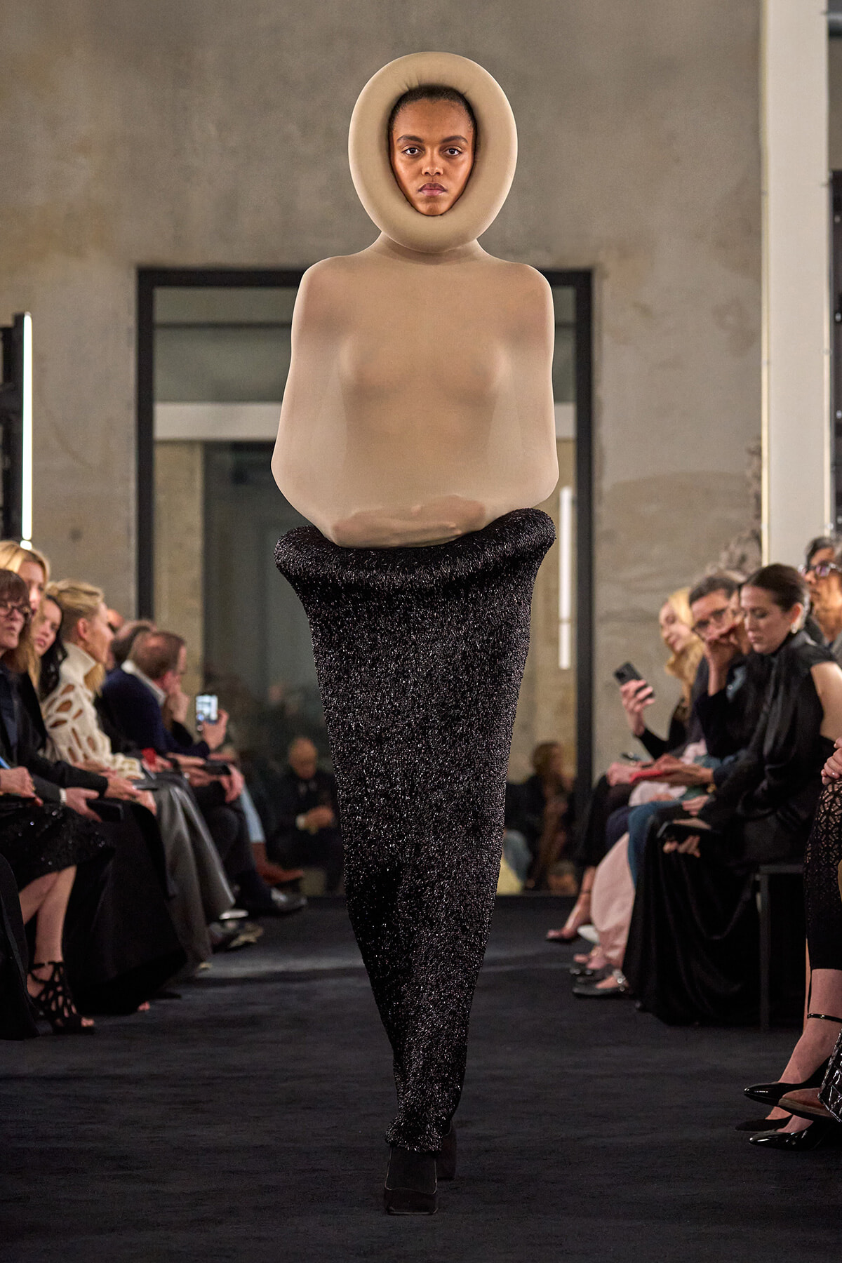 Runway model wearing a nude, sculptural bodysuit with a large circular collar around the head, paired with a glittery black bottom piece, walking the catwalk as an audience watches.