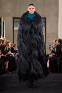 Model struts a runway in a long, dark fur coat with a bright teal scarf, audience watching and photographing on both sides.