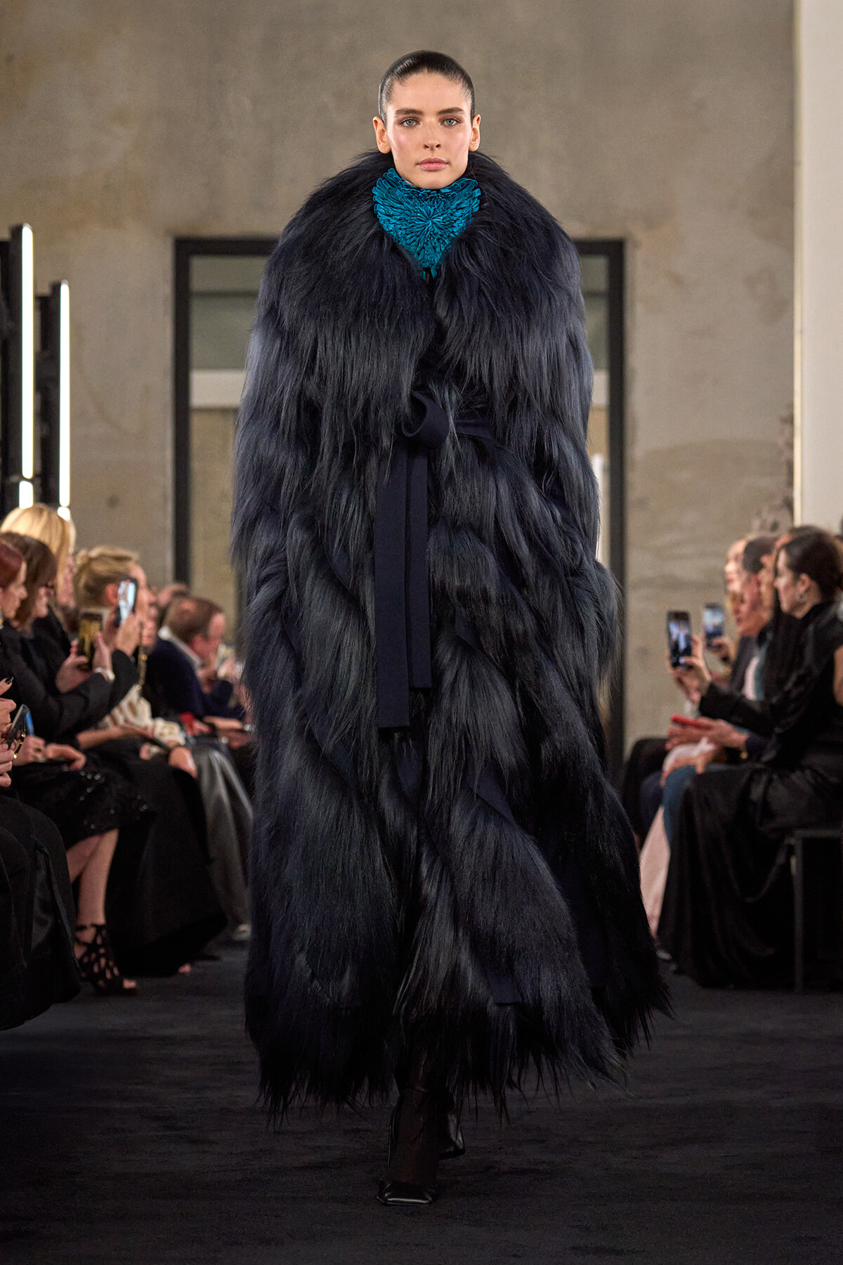 Model struts a runway in a long, dark fur coat with a bright teal scarf, audience watching and photographing on both sides.