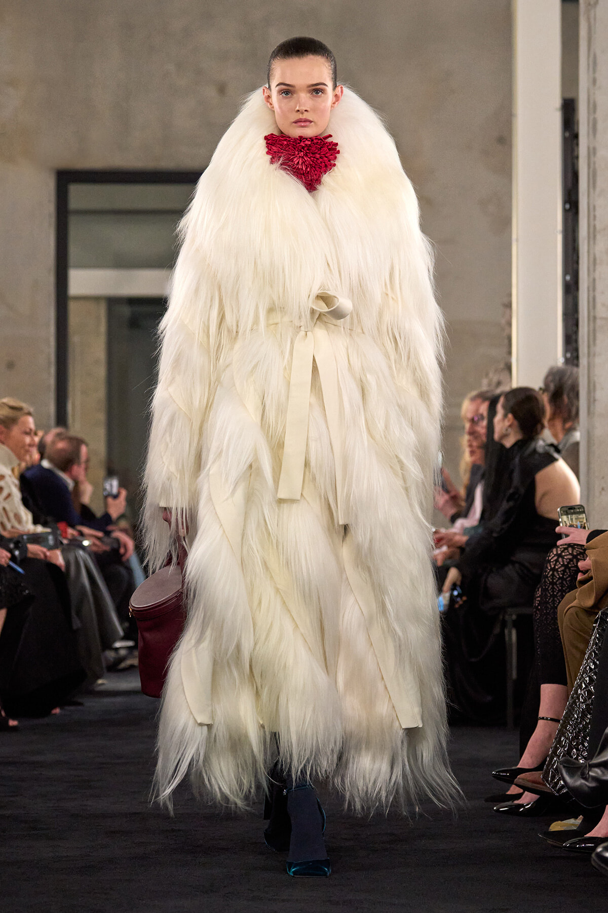 Model walking the runway in a voluminous white fur coat with a red scarf—fashion show scene with audience along the sides