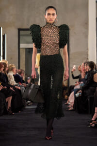 Model walks a runway in a black crochet dress with dramatic puff shoulders, carrying a structured black handbag.