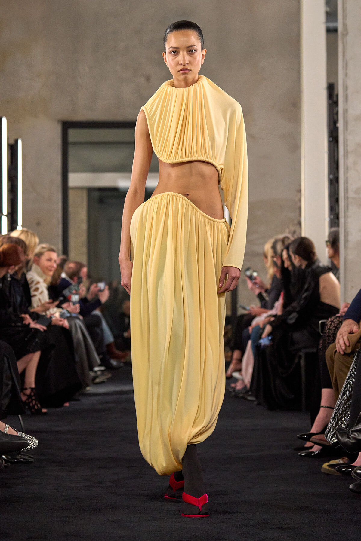 Runway model in a pale yellow draped two-piece outfit with a cutout at the waist, walking the catwalk.