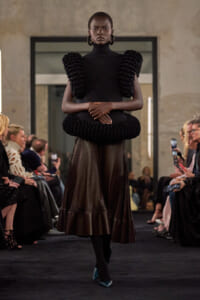 Runway model wearing a black high-neck dress with oversized shoulder ruffles and a circular waist ruffle, plus a sheer skirt and teal heels, walking the catwalk.
