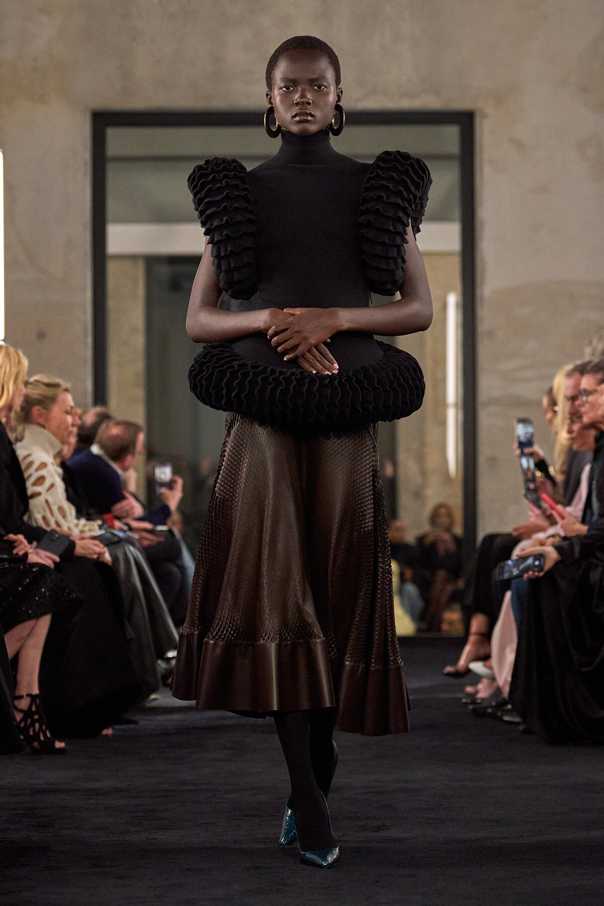 Runway model wearing a black high-neck dress with oversized shoulder ruffles and a circular waist ruffle, plus a sheer skirt and teal heels, walking the catwalk.