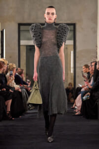 Model on a runway wearing a dark gray textured midi dress with oversized ruffled shoulders and a pale beige handbag.