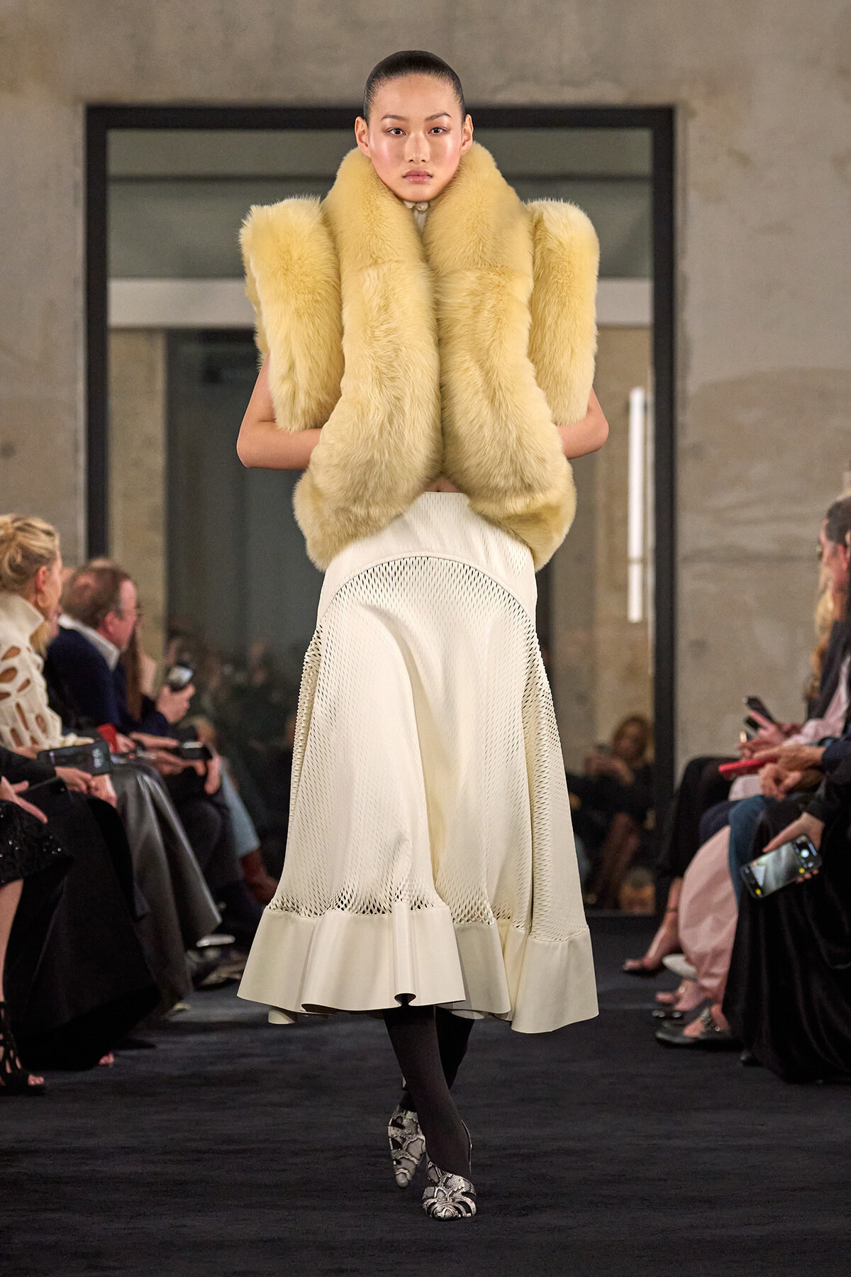 Model on a runway wearing a large cream fur vest over a white, textured midi skirt; snakeskin heels and dark tights.