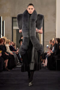 Model walks a fashion runway wearing an oversized gray fur scarf and matching fur vest, with a black skirt and high heels, audience on both sides behind.