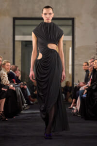 Model on a runway wearing a floor-length black draped gown with cut-out at the waist and gathered fabric at the chest, neutral expression.