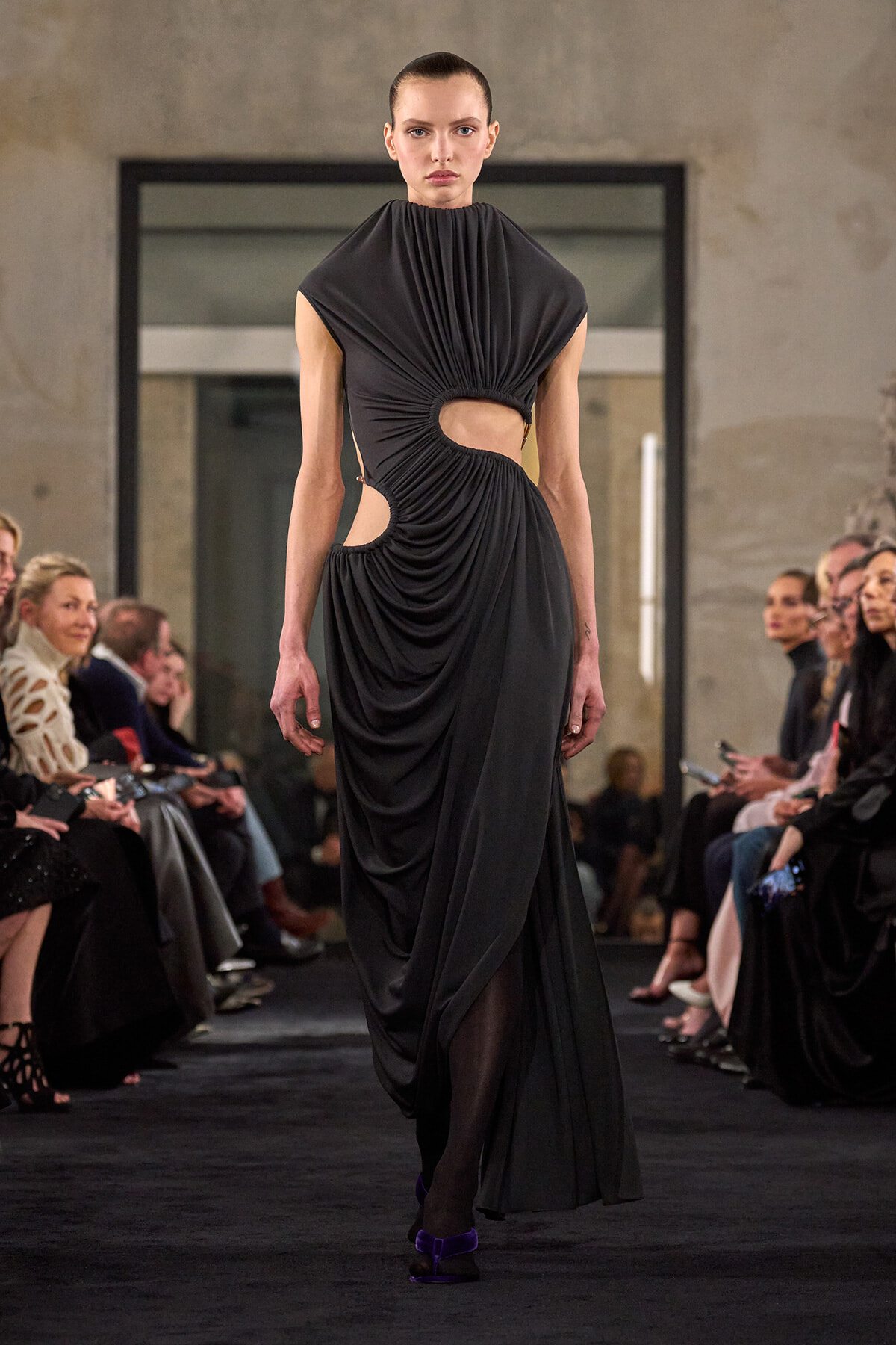 Model on a runway wearing a floor-length black draped gown with cut-out at the waist and gathered fabric at the chest, neutral expression.