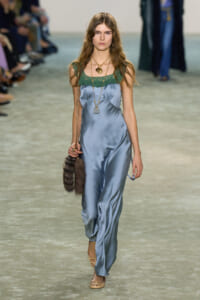 Model walking a fashion runway in a light blue satin jumpsuit with green lace neckline, layered gold necklaces, and a fur handbag with a chain strap.