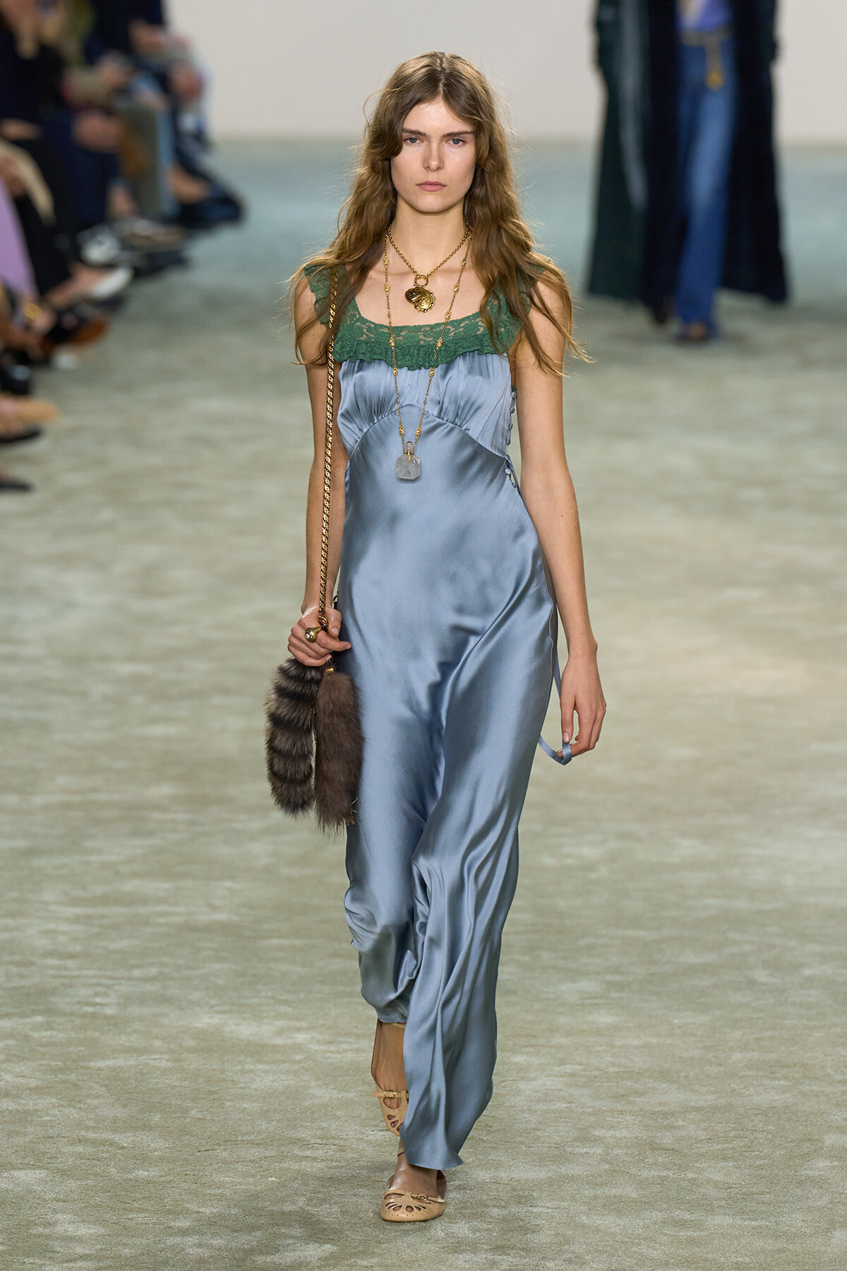Model walking a fashion runway in a light blue satin jumpsuit with green lace neckline, layered gold necklaces, and a fur handbag with a chain strap.