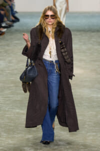 Model on a fashion runway wearing a long plaid coat with fur trim, lace blouse, blue jeans, sunglasses, and a dark handbag.