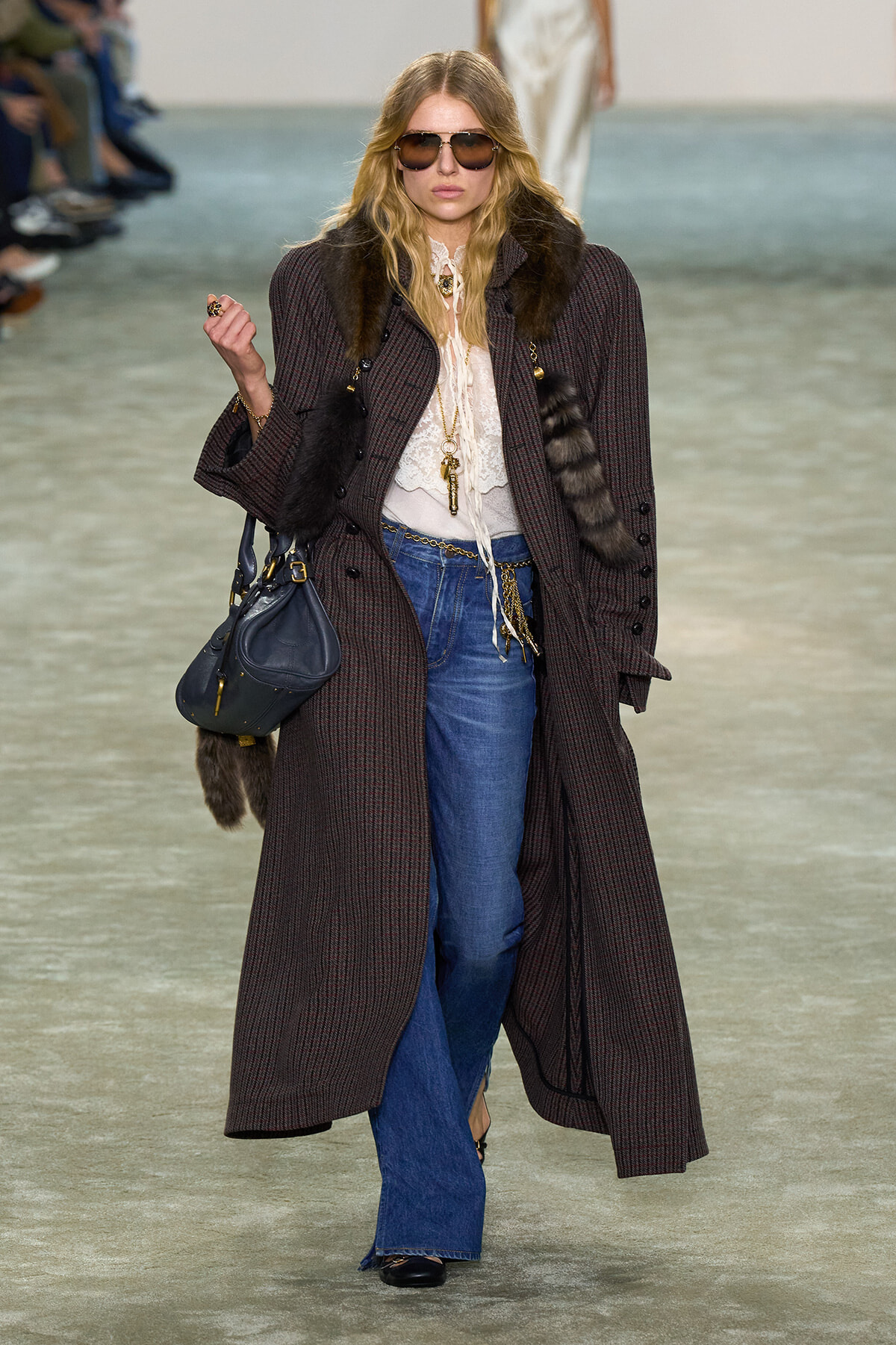 Model on a fashion runway wearing a long plaid coat with fur trim, lace blouse, blue jeans, sunglasses, and a dark handbag.