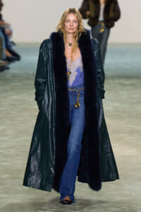 Model walking runway in a long dark coat with navy fur trim over a lavender lace top and blue jeans, accessorized with gold jewelry.
