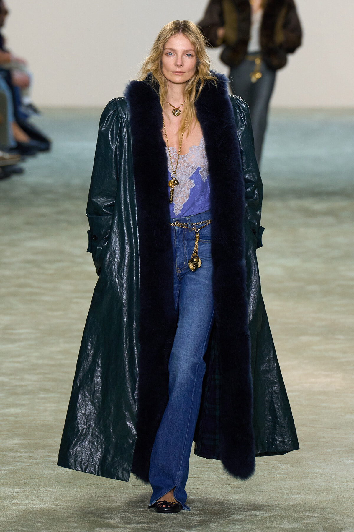 Model walking runway in a long dark coat with navy fur trim over a lavender lace top and blue jeans, accessorized with gold jewelry.