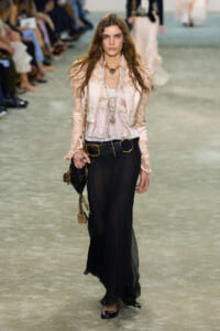 Model walking a runway in a blush pink lace blouse, cream jacket, and black sheer wide-leg pants with a gold belt and layered gold jewelry, carrying a small bag.