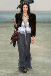 Model walks the runway in a dark fur jacket over a light gray lace blouse and flowing gray wide-leg pants, accessorized with gold jewelry and a maroon handbag.