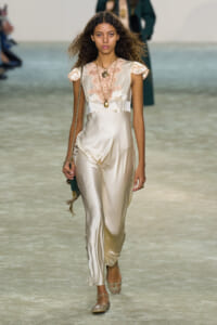 Model walking a runway in a silky cream jumpsuit with lace bodice and cap sleeves, accessorized with layered necklaces and a green handbag on a chain.