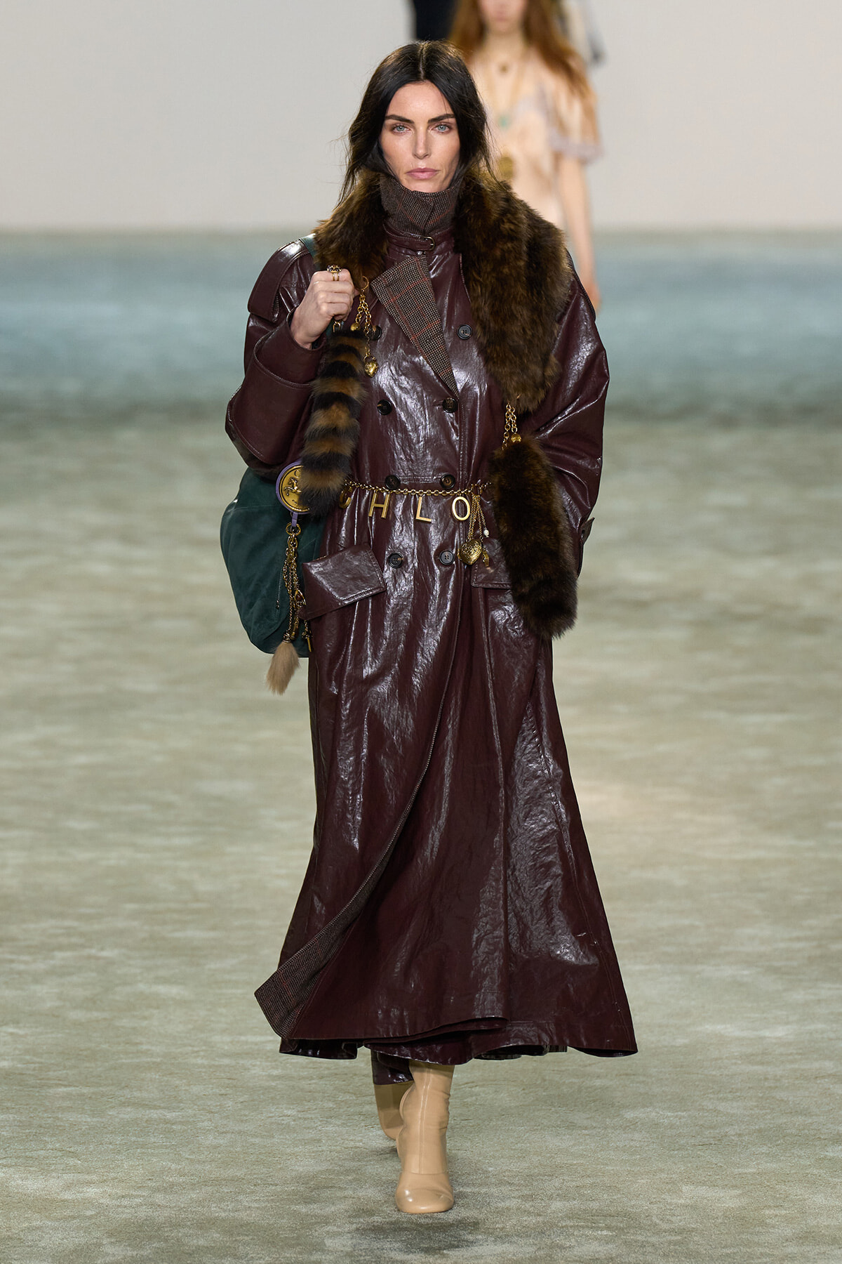 Model on a runway wearing a burgundy patent-leather trench with fur scarf and gold accessories, mid-stride down the catwalk, tan boots visible.