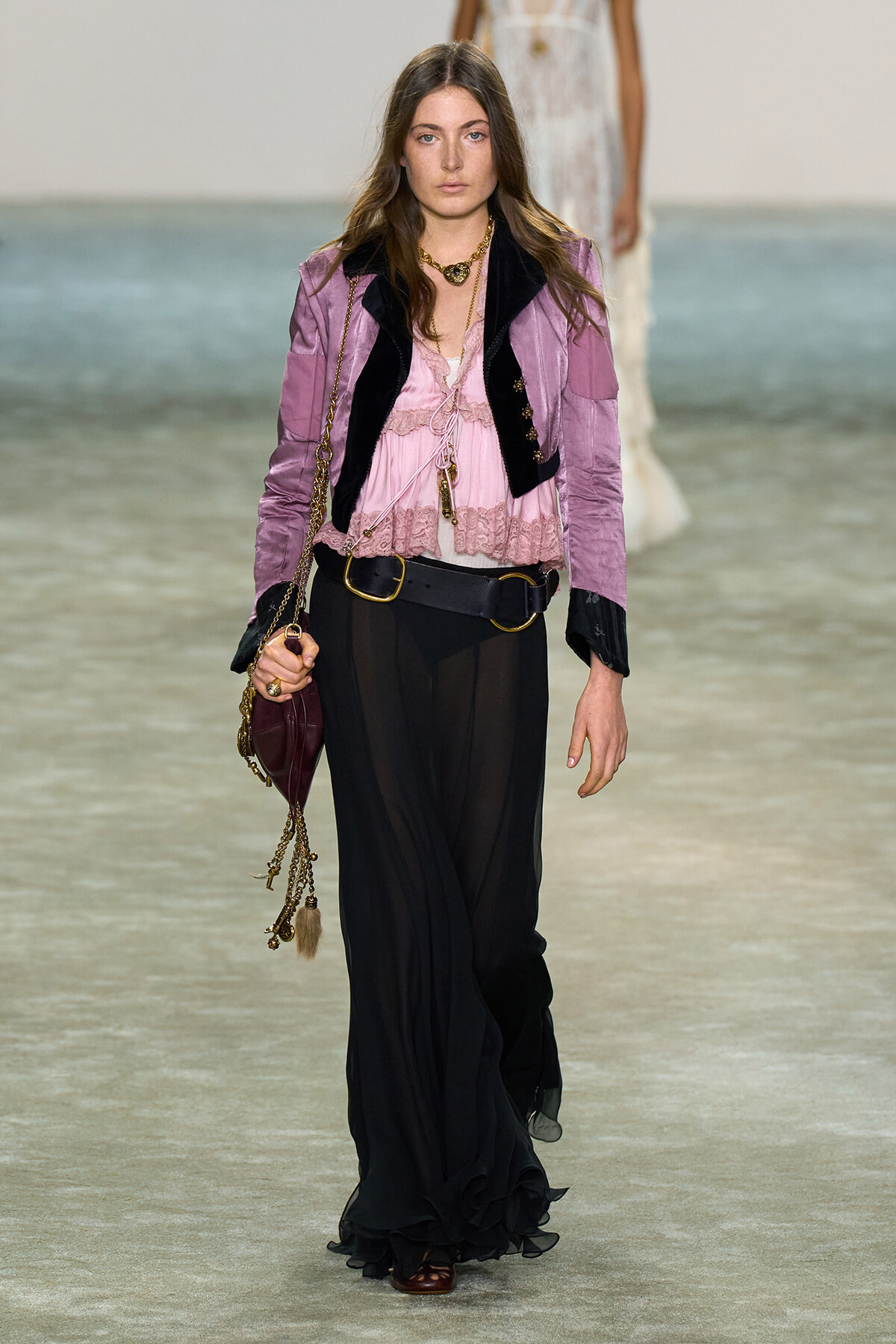 Female model walks a runway in a pink lace blouse, lavender velvet blazer, and wide black pants, carrying a burgundy bag with gold chain.