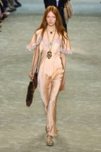 Model walks the runway in a peach satin cold-shoulder dress with lace trim, layered necklaces, and a fur-keychain bag.