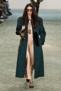 Model walking runway in a long dark green quilted coat over a beige satin jumpsuit with fur accents and large sunglasses, carrying a handbag with gold accessories.