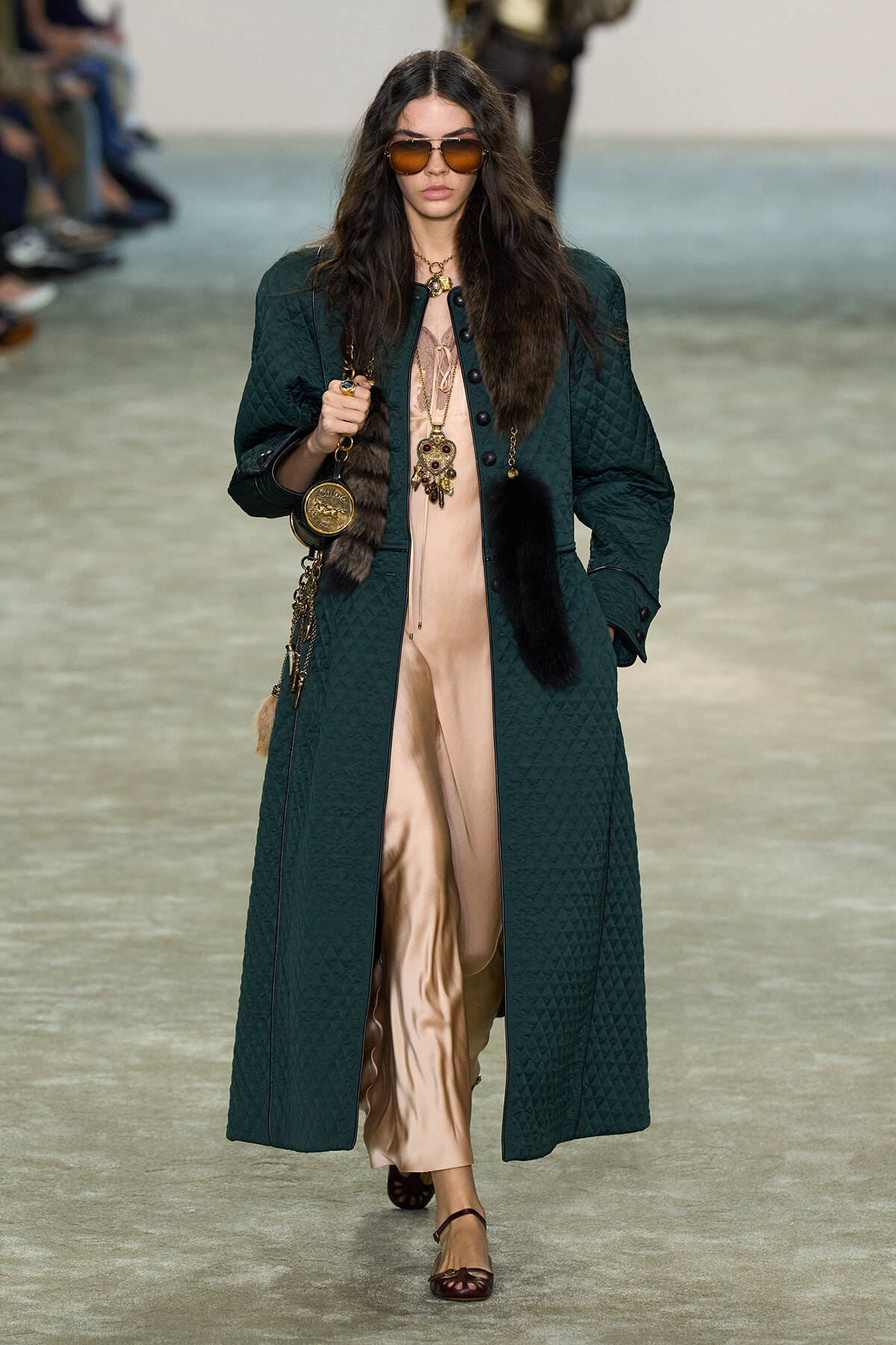 Model walking runway in a long dark green quilted coat over a beige satin jumpsuit with fur accents and large sunglasses, carrying a handbag with gold accessories.