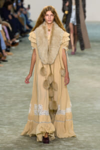 Model walking a runway in a long beige dress with a thick fur stole and tassel pom-poms; blurred audience in background.