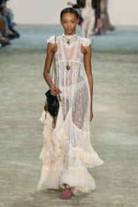 Model walking a runway in a sheer white lace gown with high neck, cap sleeves, and a layered ruffle skirt; accessorized with gold jewelry and a black handbag.