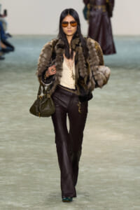 Runway model wearing a brown fur coat, beige top, dark leather pants, orange sunglasses, and carrying an olive handbag.