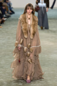 Runway model walks wearing a taupe tiered chiffon gown with a fluffy fur stole and dangling gold accessories.