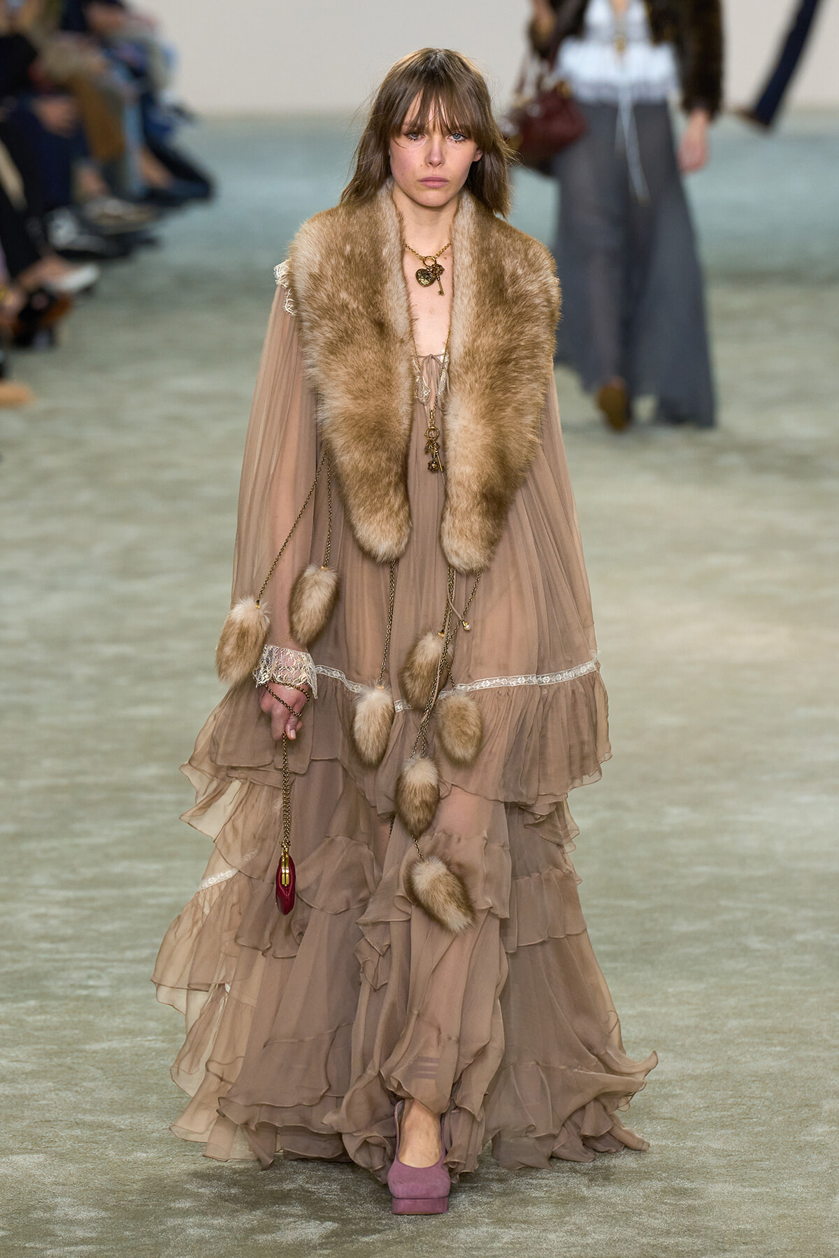 Runway model walks wearing a taupe tiered chiffon gown with a fluffy fur stole and dangling gold accessories.
