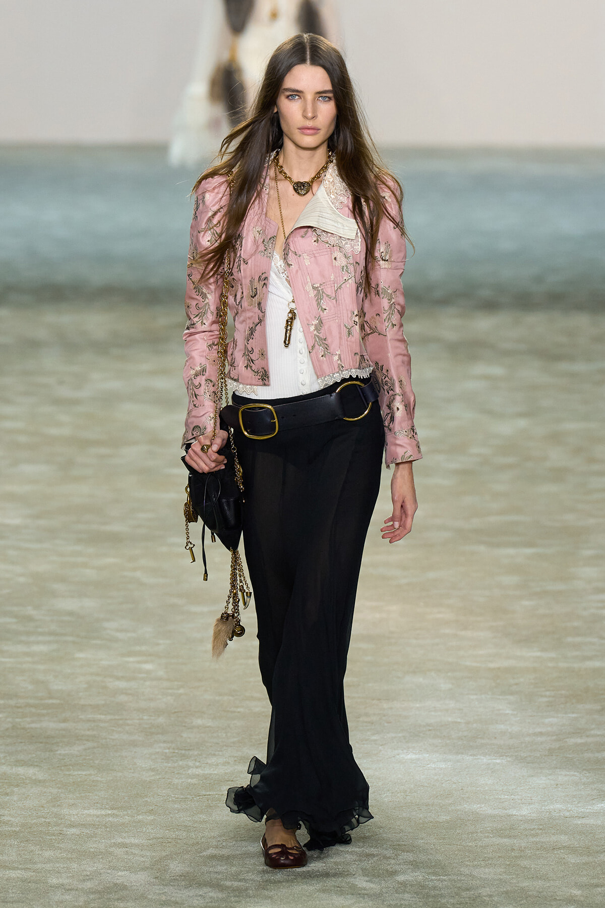 Model walking runway in a pink embroidered jacket over a white top, paired with black wide-leg pants and a gold-buckle belt, carrying a chained handbag.