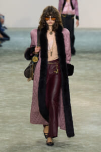 Model walks the runway in a long pink quilted coat with black fur trim, burgundy leather pants, and layered gold accessories, wearing brown sunglasses.