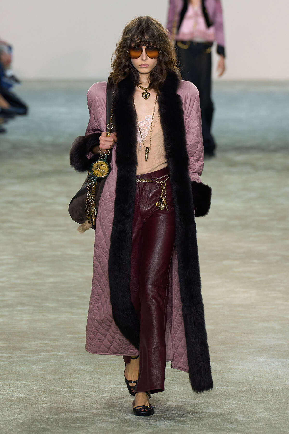 Model walks the runway in a long pink quilted coat with black fur trim, burgundy leather pants, and layered gold accessories, wearing brown sunglasses.